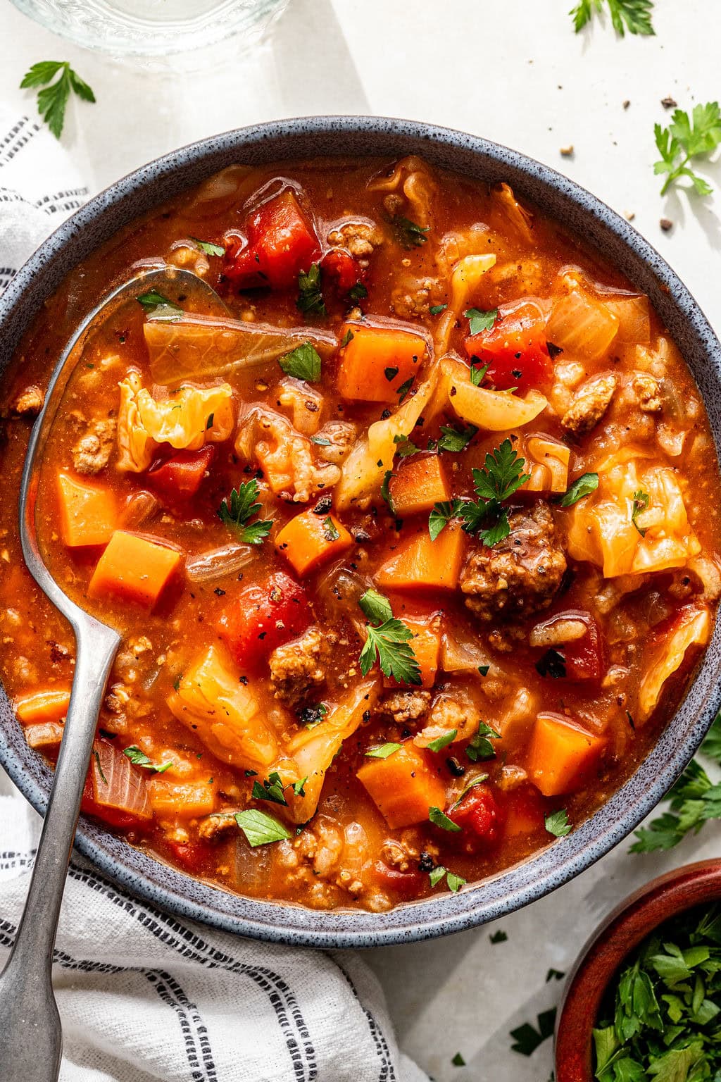 A bowl of chunky soup with ground meat, tomatoes, carrots, cabbage, and herbs, with a spoon inside the bowl and fresh parsley garnished on top.