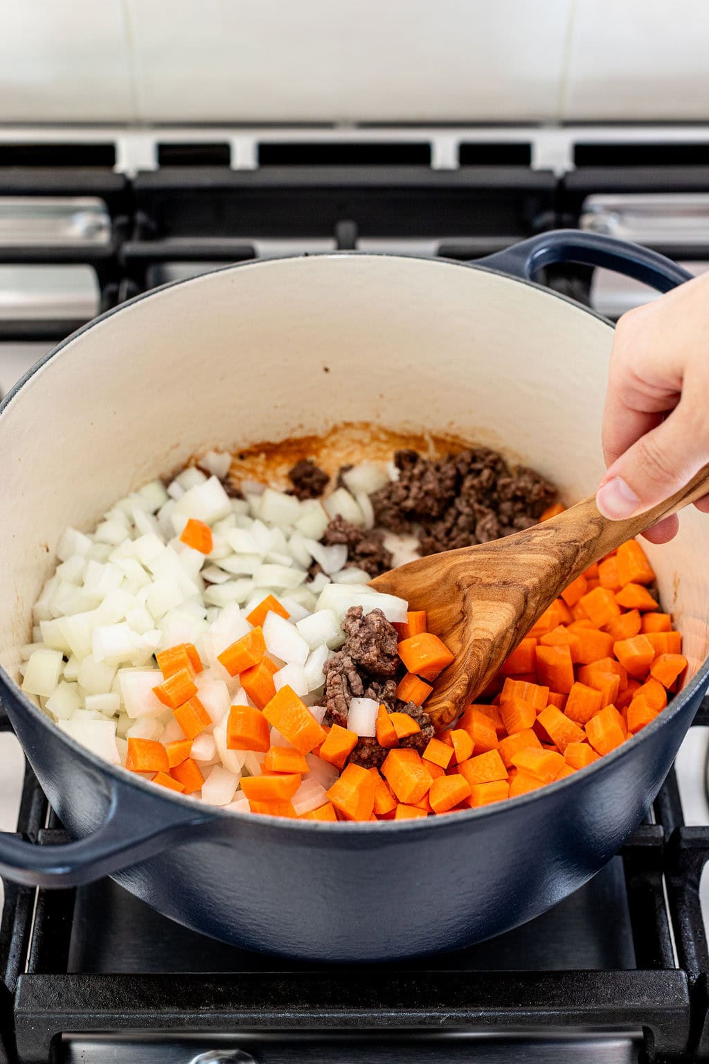 A hand stirs diced onions, carrots, and cooked ground beef in a large blue pot on a stovetop.