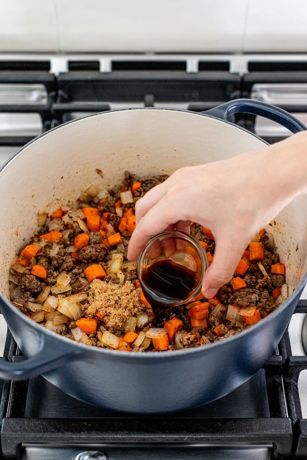 A hand pours a small glass of Worcestershire sauce into a pot containing ground meat, chopped onions, carrots, and brown sugar on a stovetop.