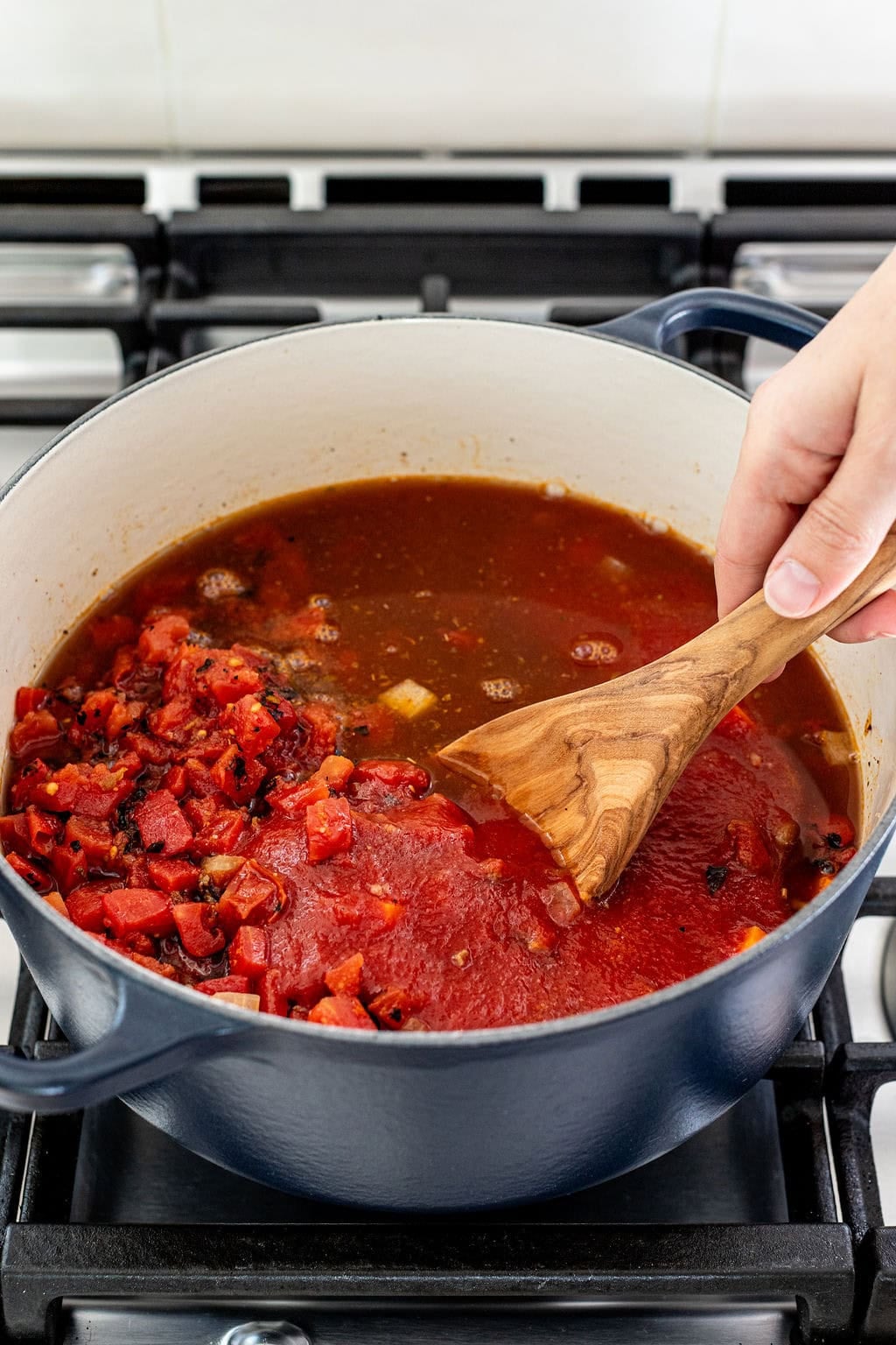 A hand stirs a pot of tomato sauce with a wooden spoon on a stovetop.