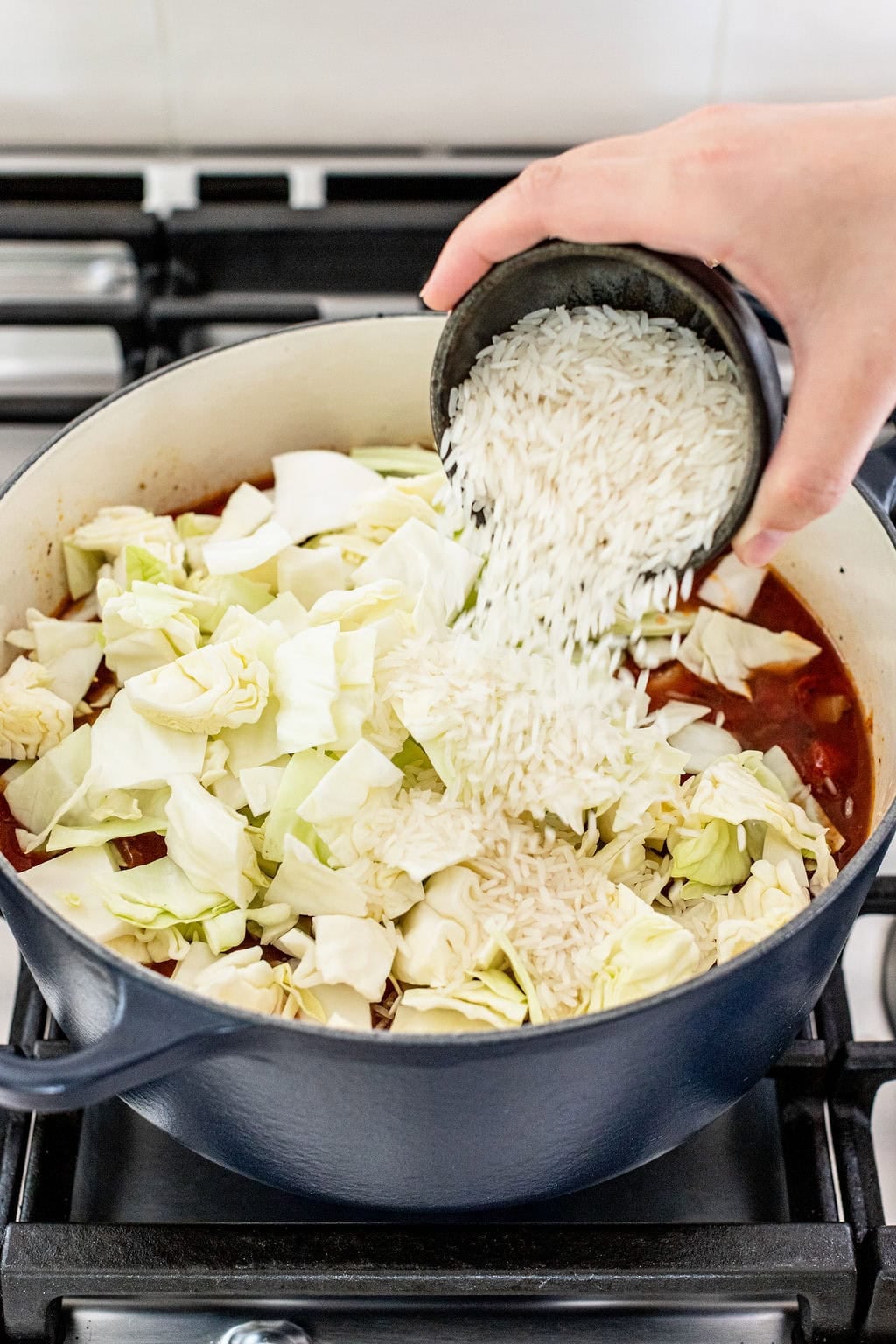 A hand pours uncooked white rice into a pot containing chopped cabbage and a tomato-based liquid on a stovetop.