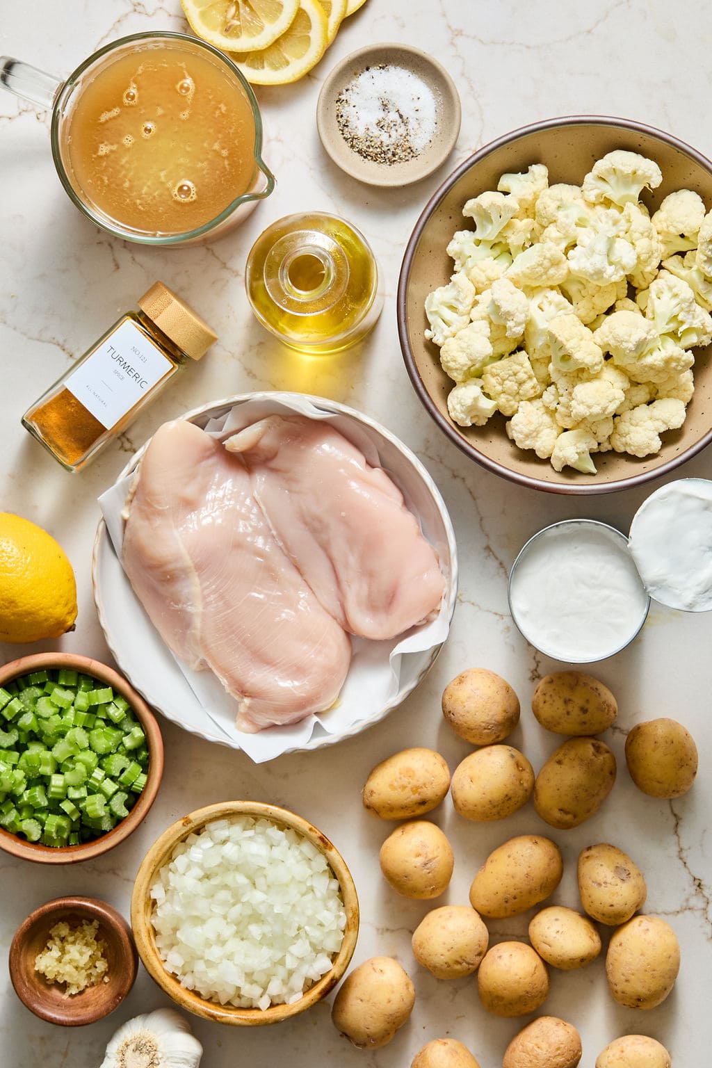 Raw chicken breasts, cauliflower, baby potatoes, celery, onion, garlic, turmeric, broth, olive oil, lemon, salt, pepper, and coconut milk are arranged on a light countertop.