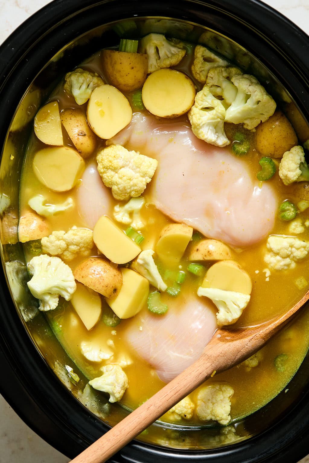 Uncooked chicken breasts, chopped potatoes, cauliflower, and celery in a slow cooker with yellow broth, stirred by a wooden spoon.