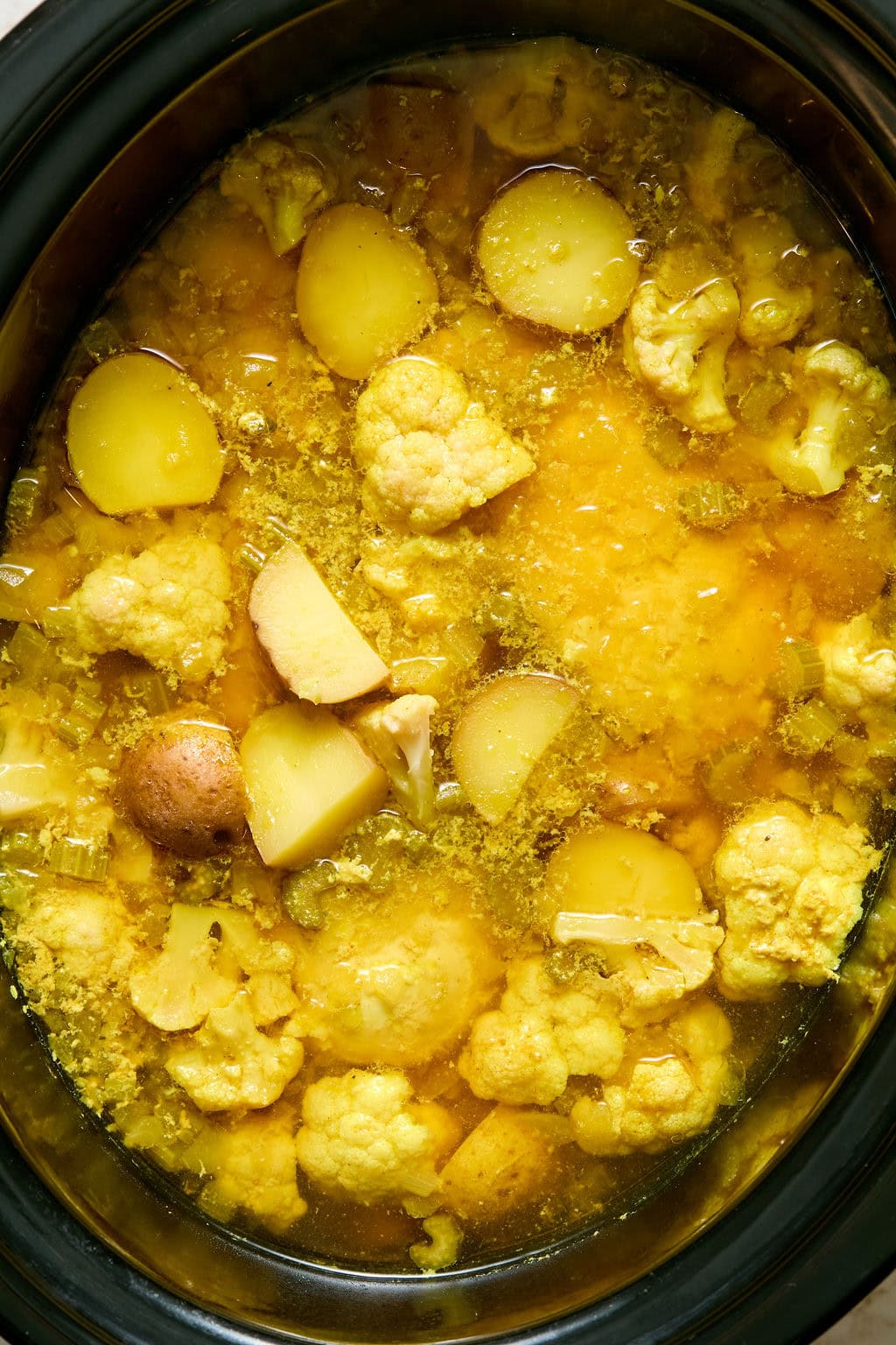 Slow cooker filled with yellow curry broth, cauliflower florets, and potato chunks.
