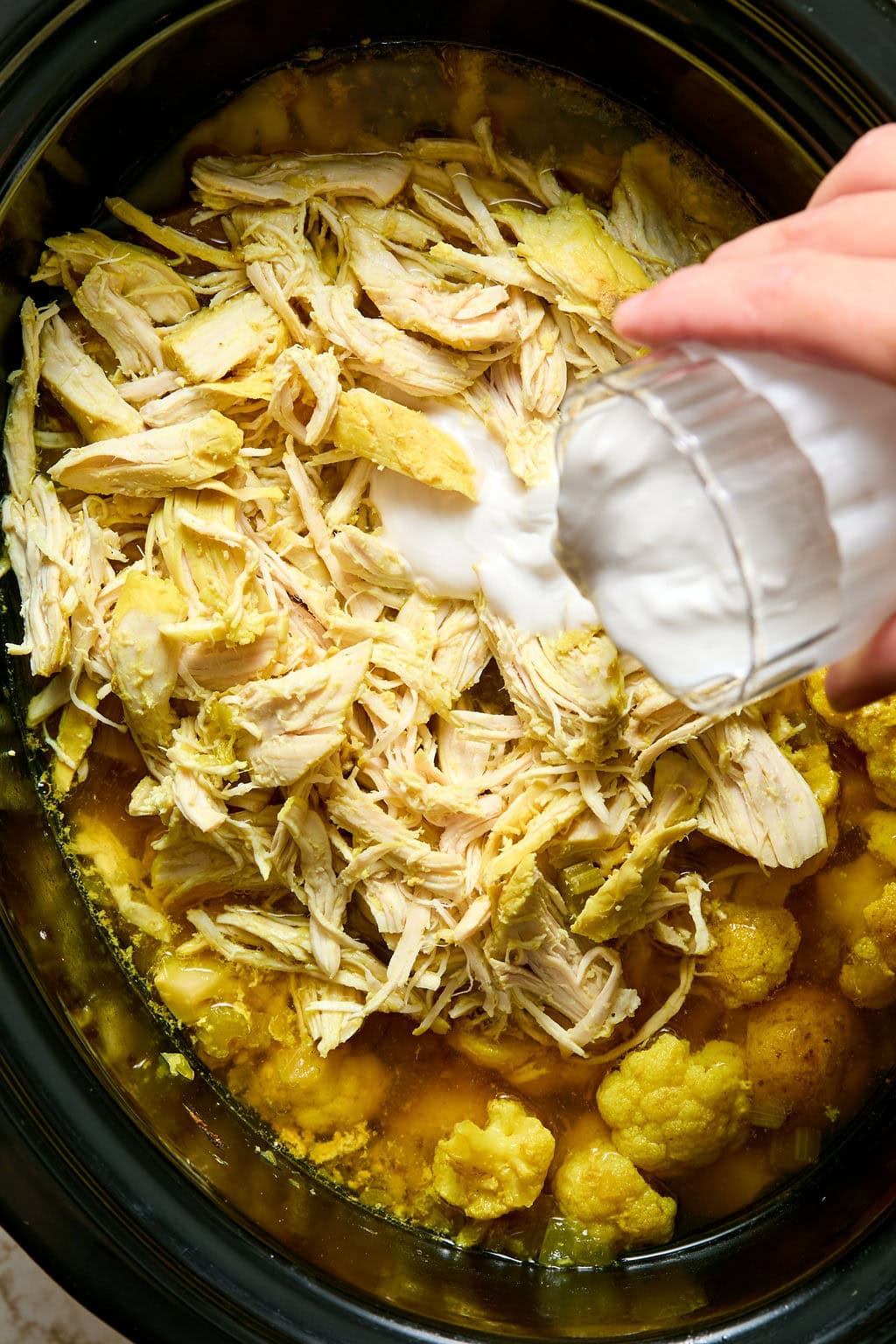 Shredded chicken and cauliflower in a slow cooker, with a hand pouring coconut milk from a glass over the mixture.