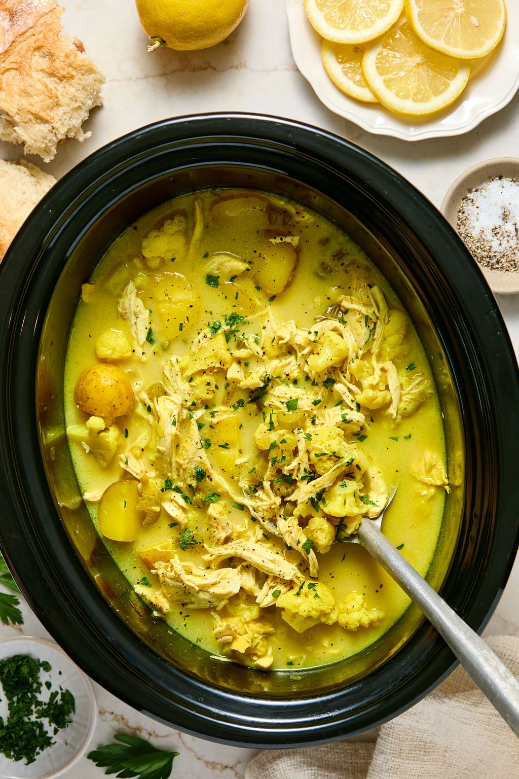 A slow cooker filled with yellow chicken stew, potatoes, and herbs, with a serving spoon. Lemon slices, bread, and seasonings are arranged around the dish.