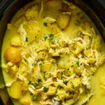 A close-up of a slow cooker filled with yellow curry chicken soup containing shredded chicken, potatoes, and herbs.
