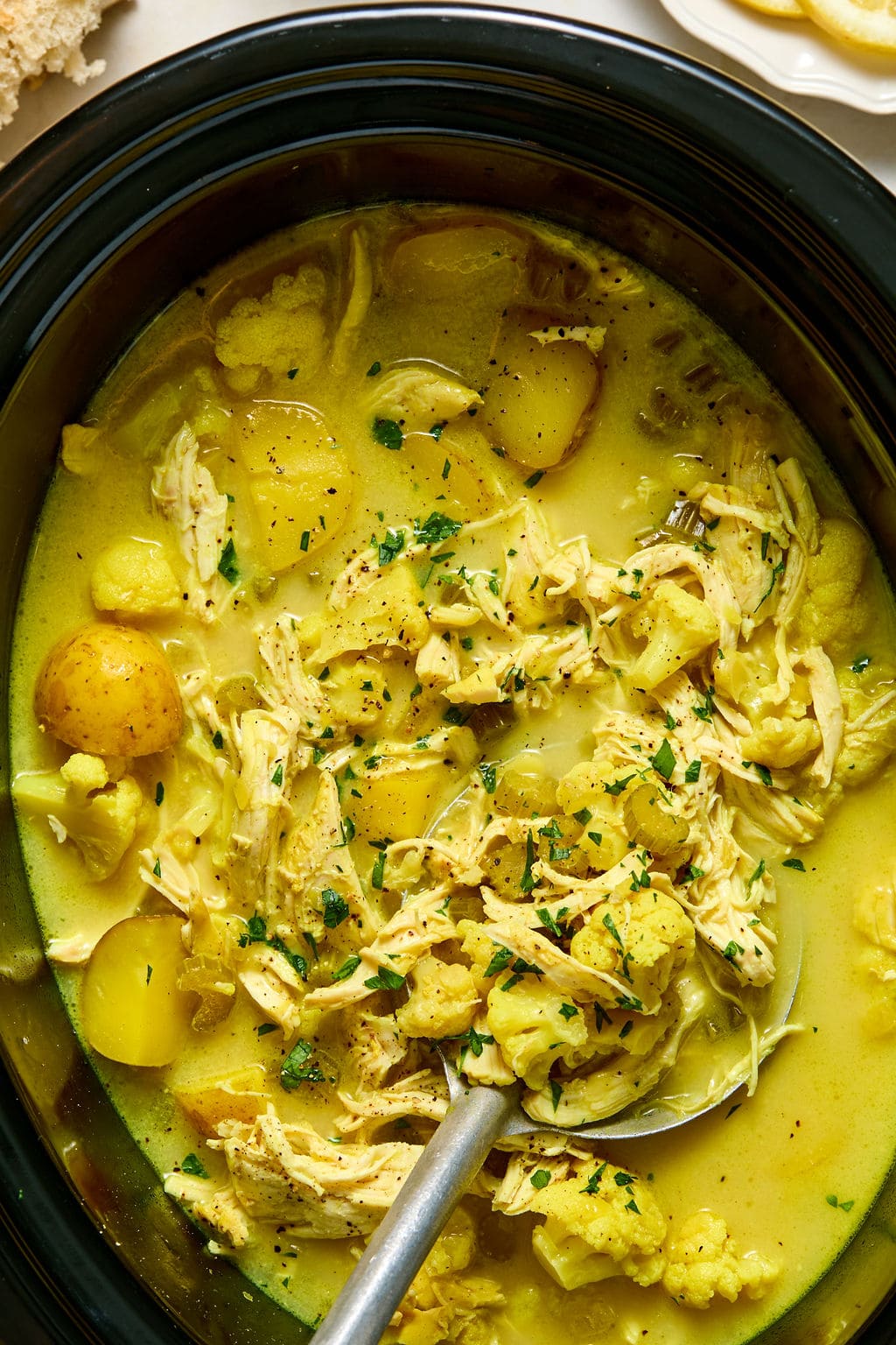 A close-up of a slow cooker filled with yellow curry chicken soup containing shredded chicken, potatoes, and herbs.