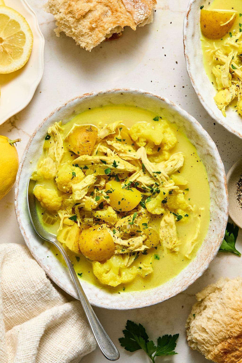 A bowl of yellow chicken and potato stew garnished with herbs, with a spoon in the bowl. Bread, lemon, and parsley are placed nearby on a light surface.
