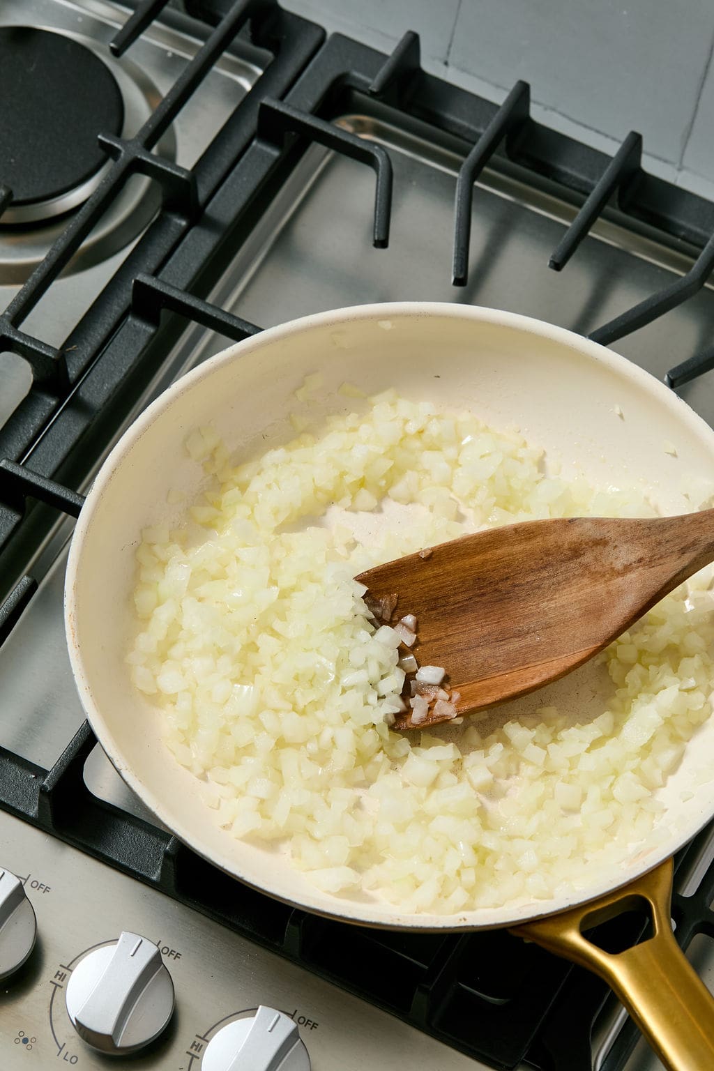 Chopped onions being saut&eacute;ed in a white frying pan on a gas stove, stirred with a wooden spatula.
