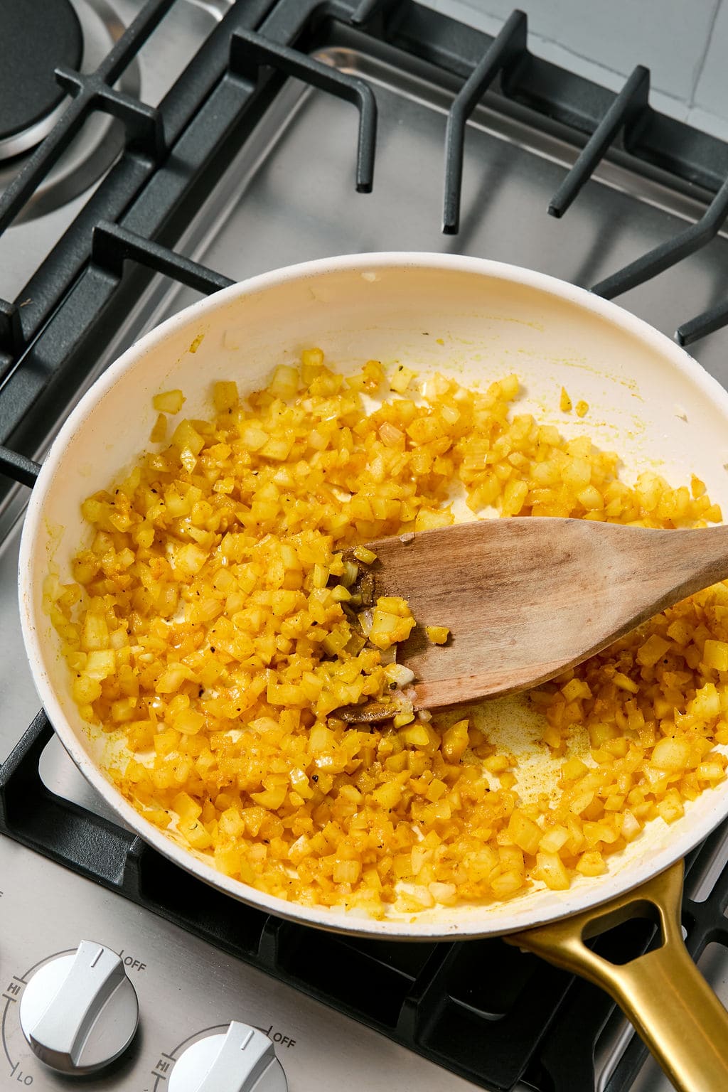Chopped onions being saut&eacute;ed in a white skillet on a stove, stirred with a wooden spatula.