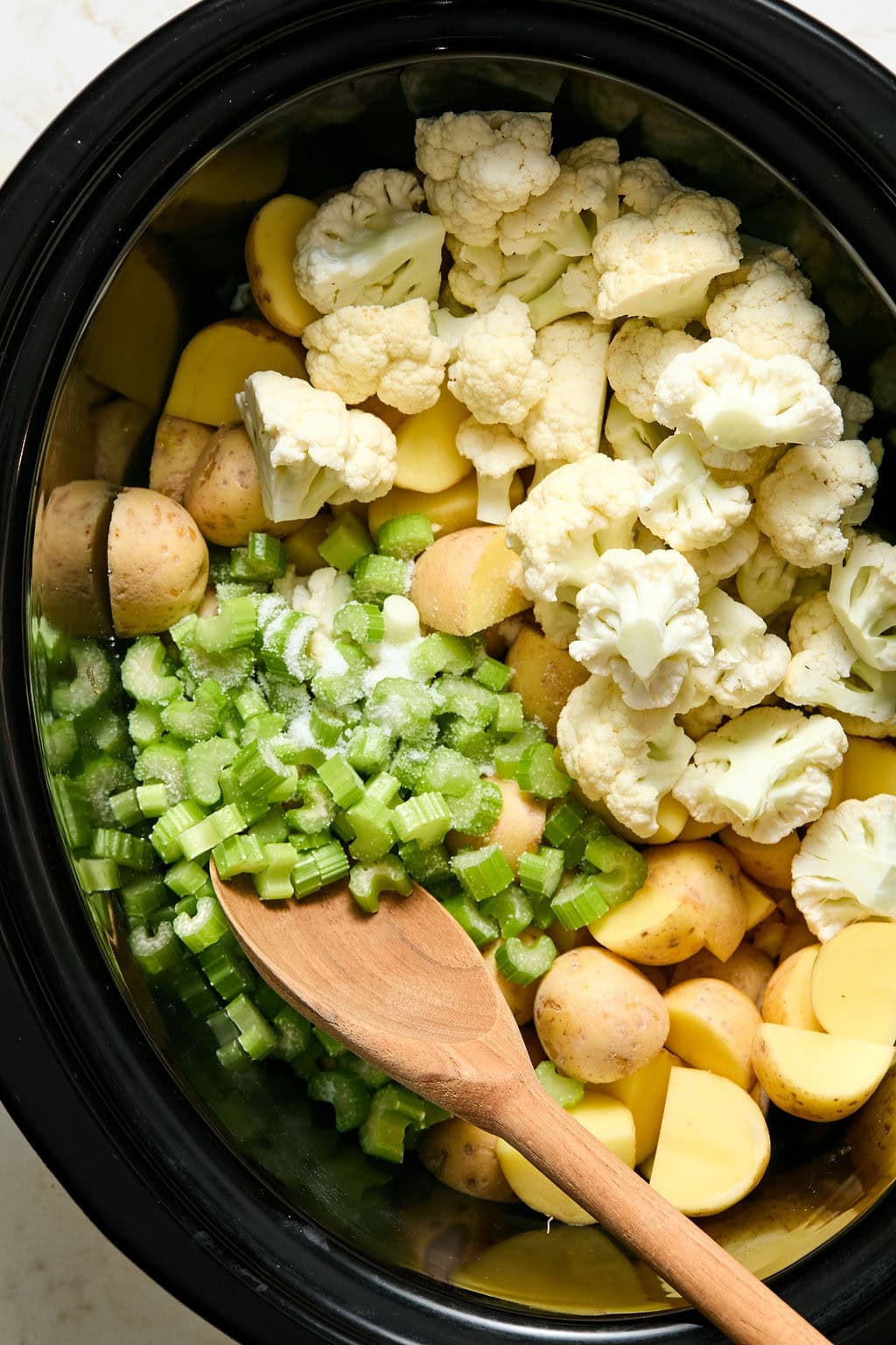 Chopped potatoes, cauliflower florets, and sliced celery in a slow cooker with a wooden spoon.