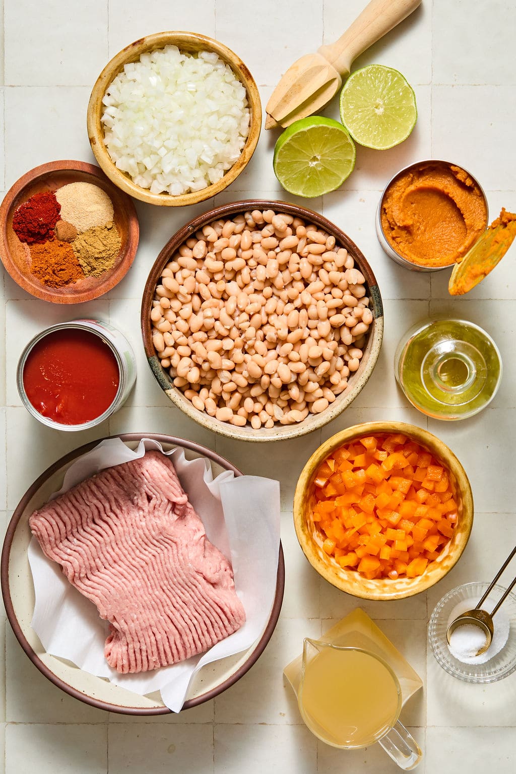 Overhead view of raw ground meat, white beans, chopped onions, bell peppers, tomato sauce, lime halves, pumpkin, broth, spices, and olive oil arranged on a light surface.