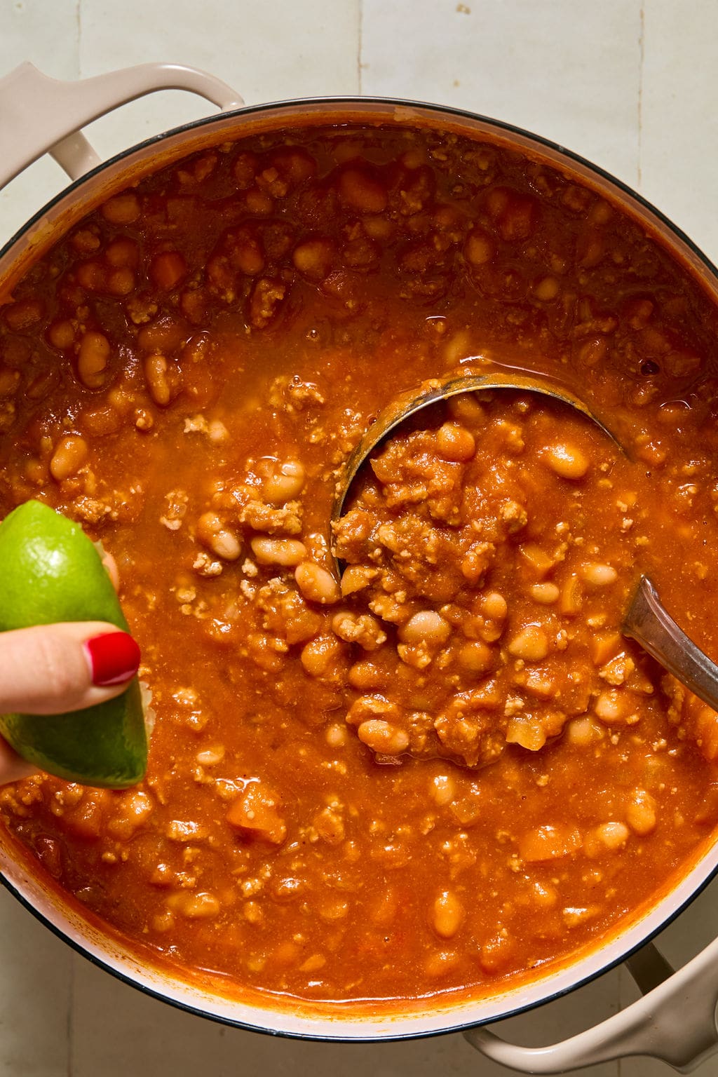 A pot of chili with beans and ground meat sits on a stovetop. A hand with red nail polish holds a lime wedge above the pot, ready to squeeze. A ladle rests in the chili.