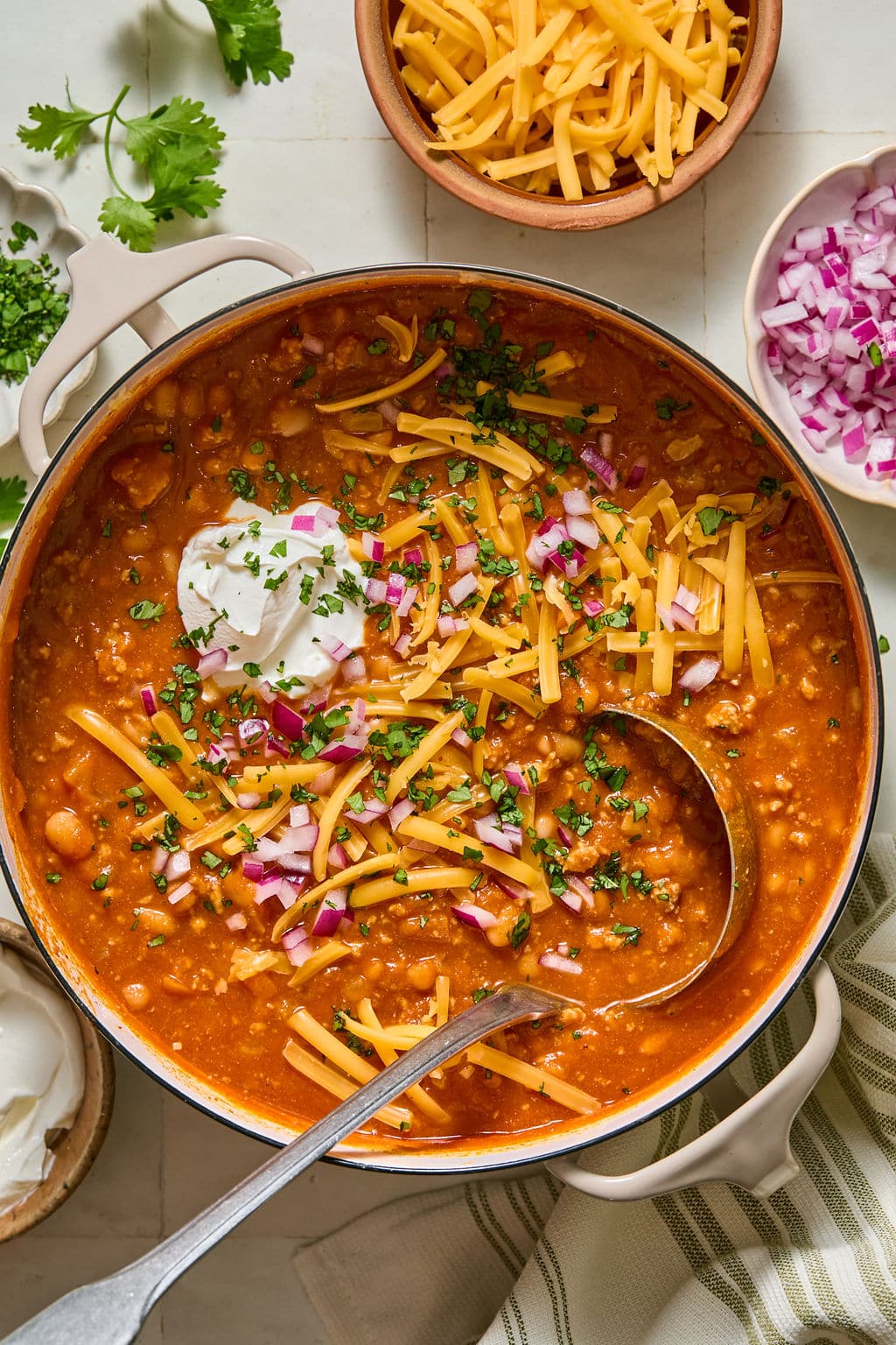 A pot of chili topped with shredded cheddar, diced red onions, chopped herbs, and a dollop of sour cream, surrounded by bowls of extra cheese, onions, and sour cream.