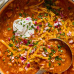 A pot of chili topped with shredded cheddar cheese, chopped red onions, fresh herbs, and a dollop of sour cream, with a metal ladle resting in the chili.