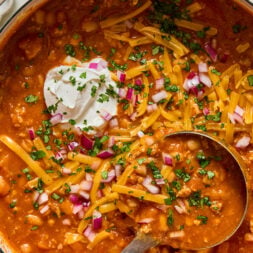 A pot of chili topped with shredded cheddar cheese, chopped red onions, fresh herbs, and a dollop of sour cream, with a metal ladle resting in the chili.