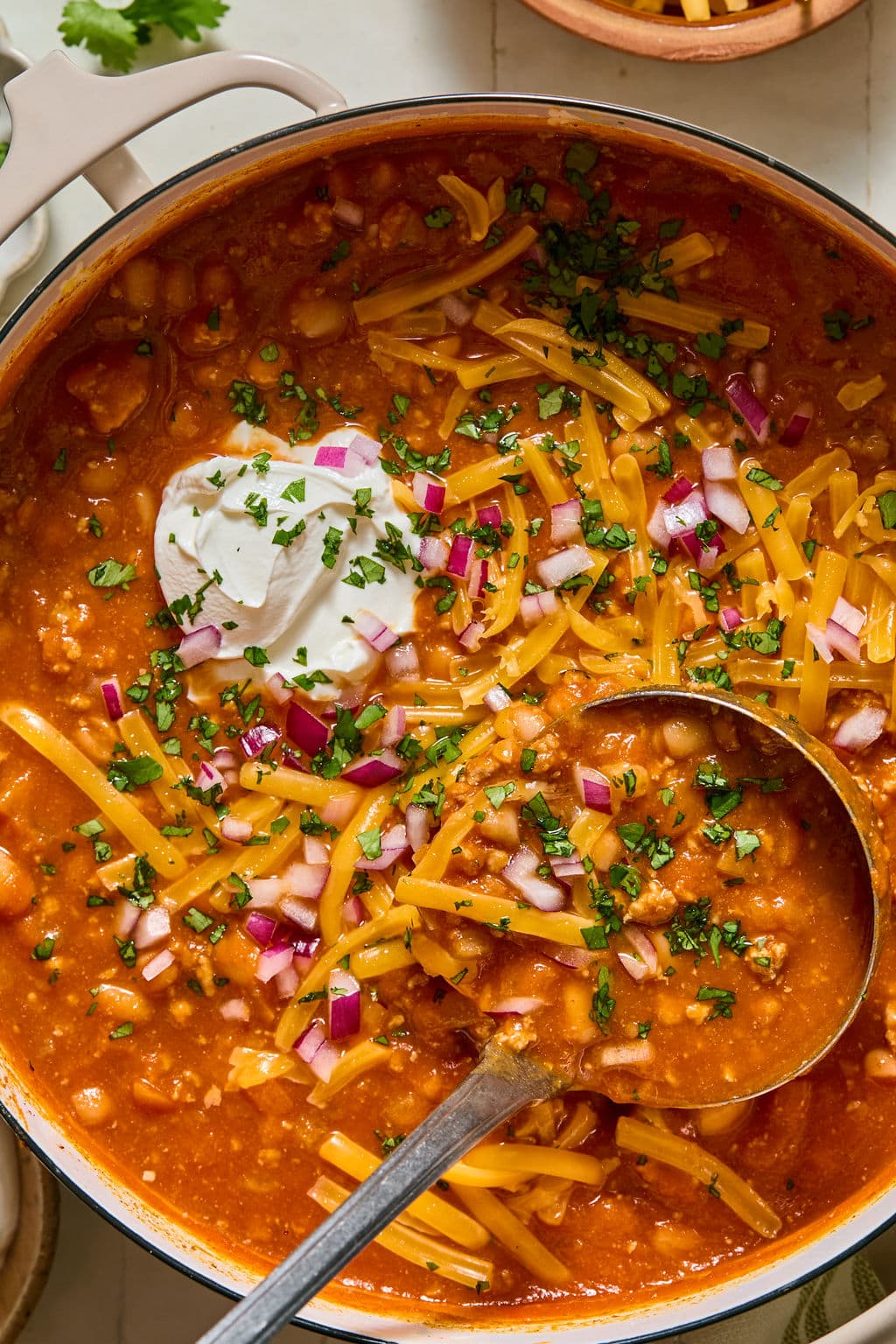 A pot of chili topped with shredded cheddar cheese, chopped red onions, fresh herbs, and a dollop of sour cream, with a metal ladle resting in the chili.