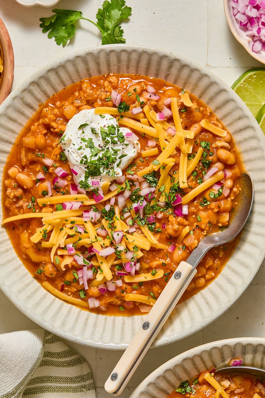 A bowl of chili topped with shredded cheese, sour cream, chopped red onions, and herbs, with a spoon resting inside the bowl.