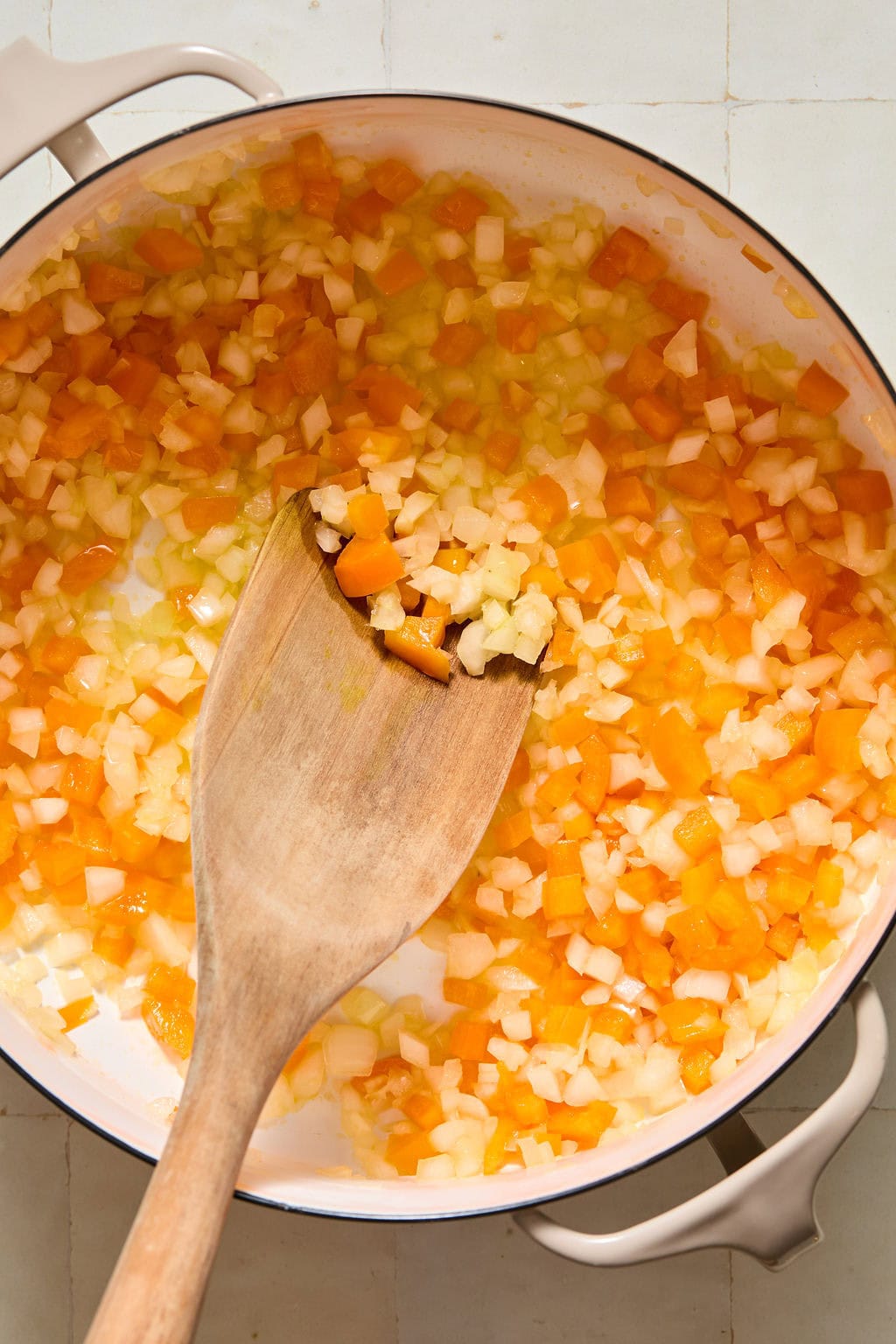 Diced onions, carrots, and celery being sautéed in a white pan with a wooden spoon on a tiled countertop.