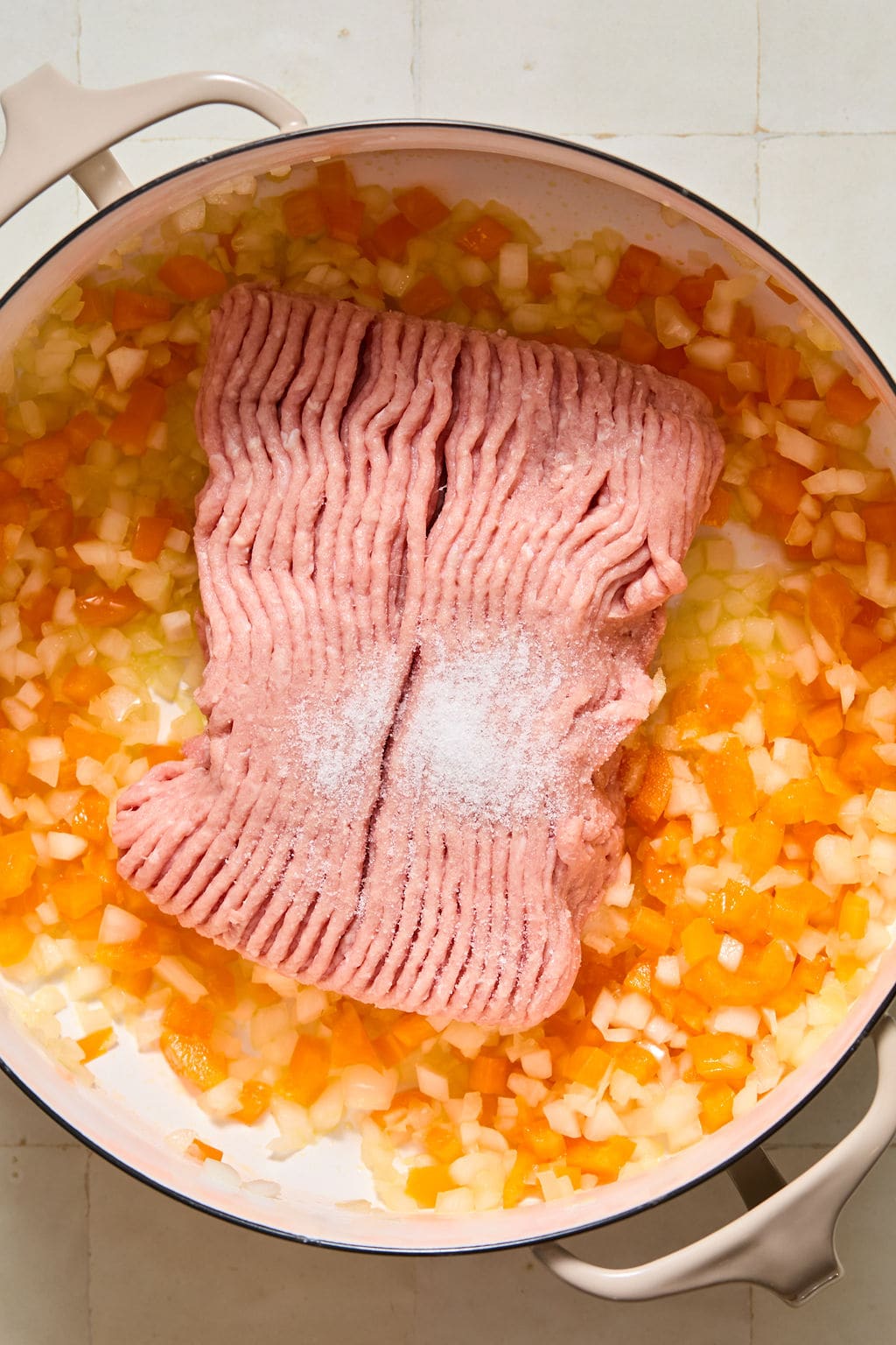 Ground turkey with salt on top sits in a pot with chopped onions and orange bell peppers on a white tiled surface.