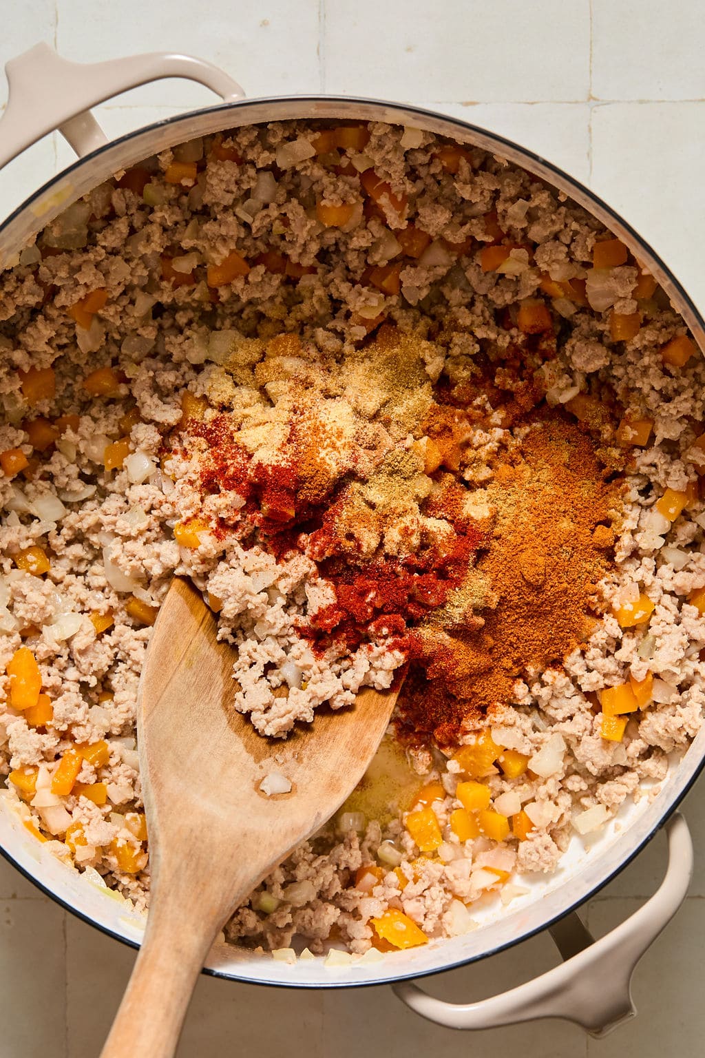Overhead view of ground meat, diced onions, and bell peppers cooking in a pot, with a wooden spoon stirring in a pile of colorful spices.