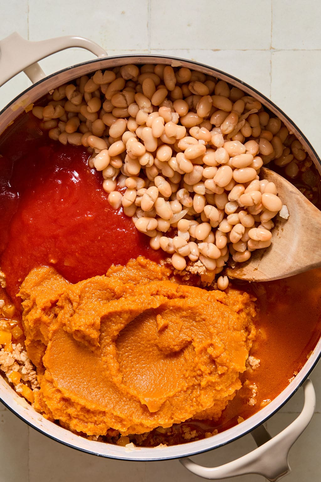 A pot containing white beans, tomato sauce, and pumpkin puree, with a wooden spoon resting inside.