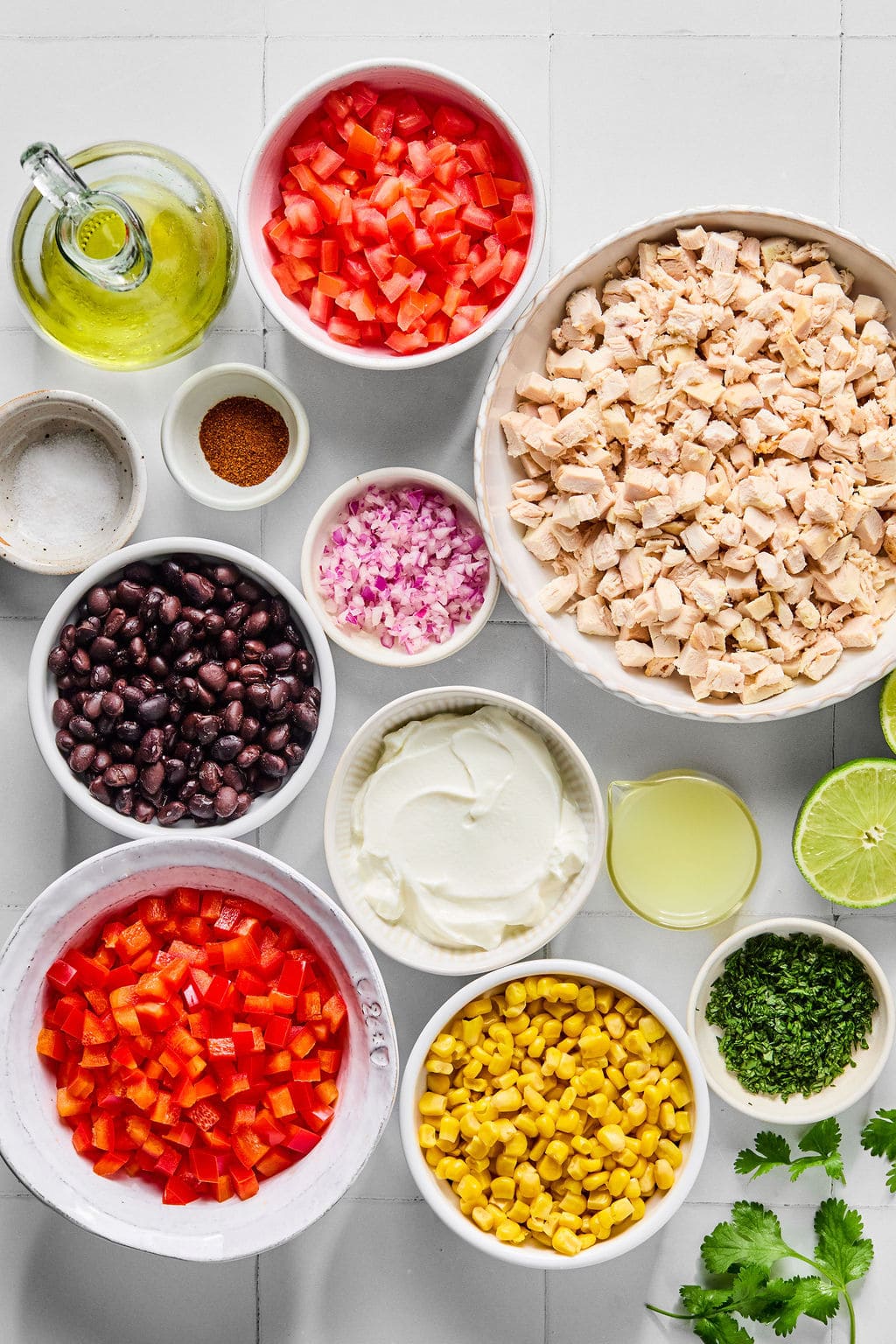 Top-down view of bowls containing diced chicken, black beans, corn, red bell pepper, tomato, red onion, sour cream, chopped herbs, lime, olive oil, spices, salt, and cilantro on a white surface.