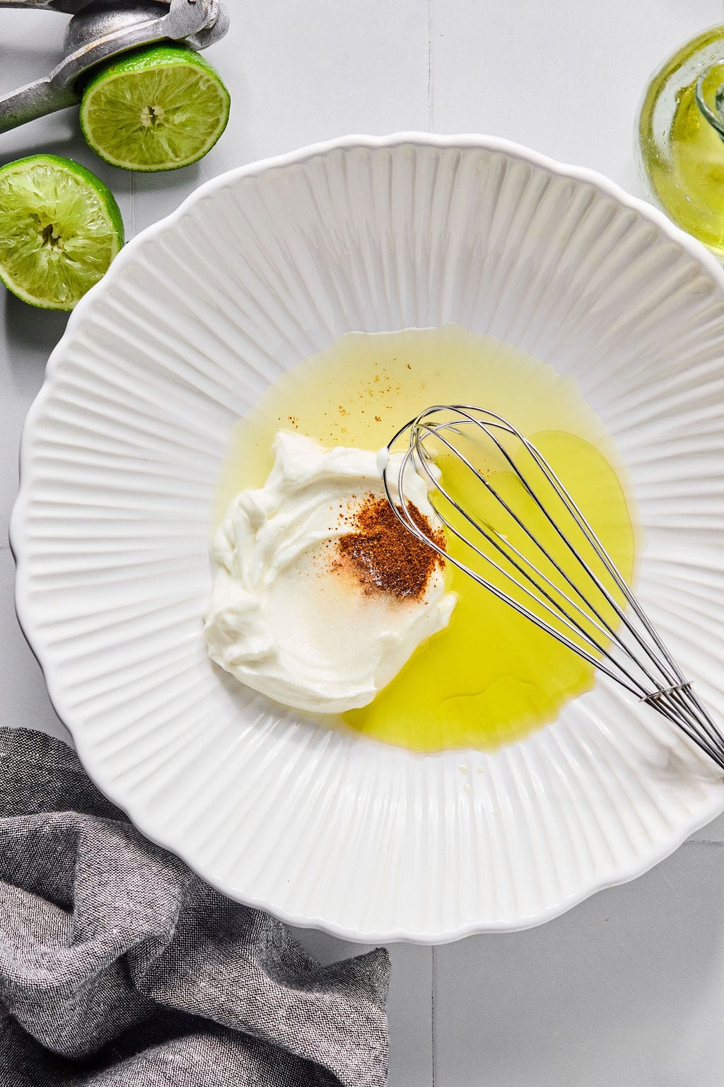 A white bowl with yogurt, olive oil, spices, and a whisk. Sliced limes, a juicer, and a gray cloth are nearby on a light surface.