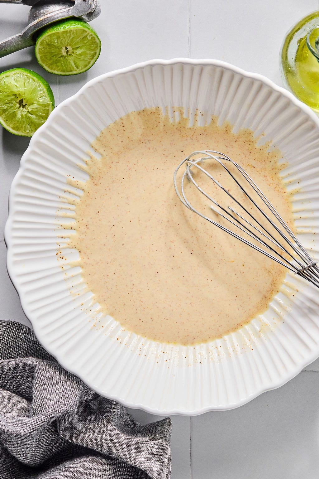 A white bowl with a creamy, speckled dressing being whisked, next to a halved lime, a lime press, and a gray cloth on a white surface.