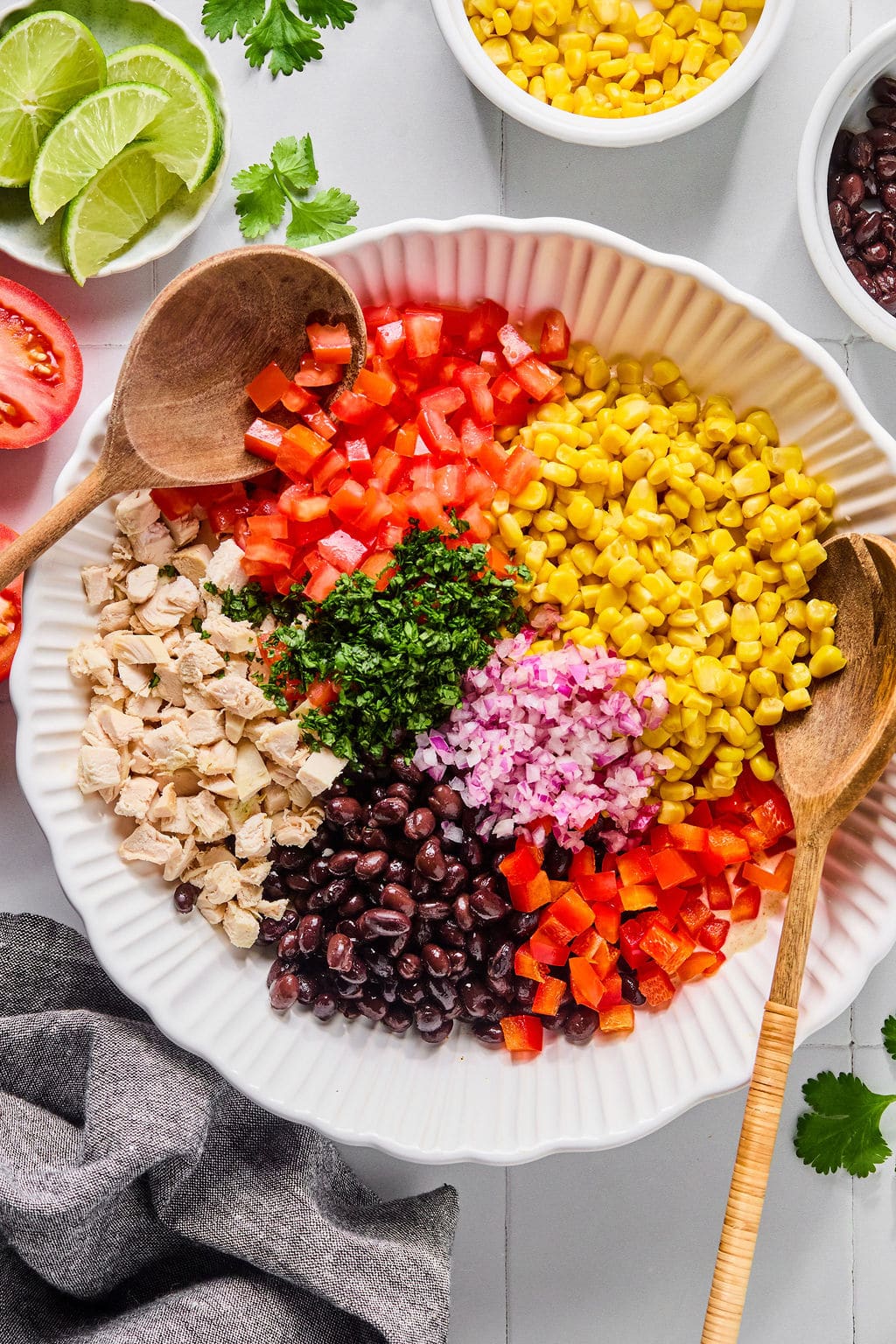 A white bowl contains chopped tomato, corn, black beans, red onion, red bell pepper, cooked chicken, and cilantro, with wooden spoons resting on the sides.