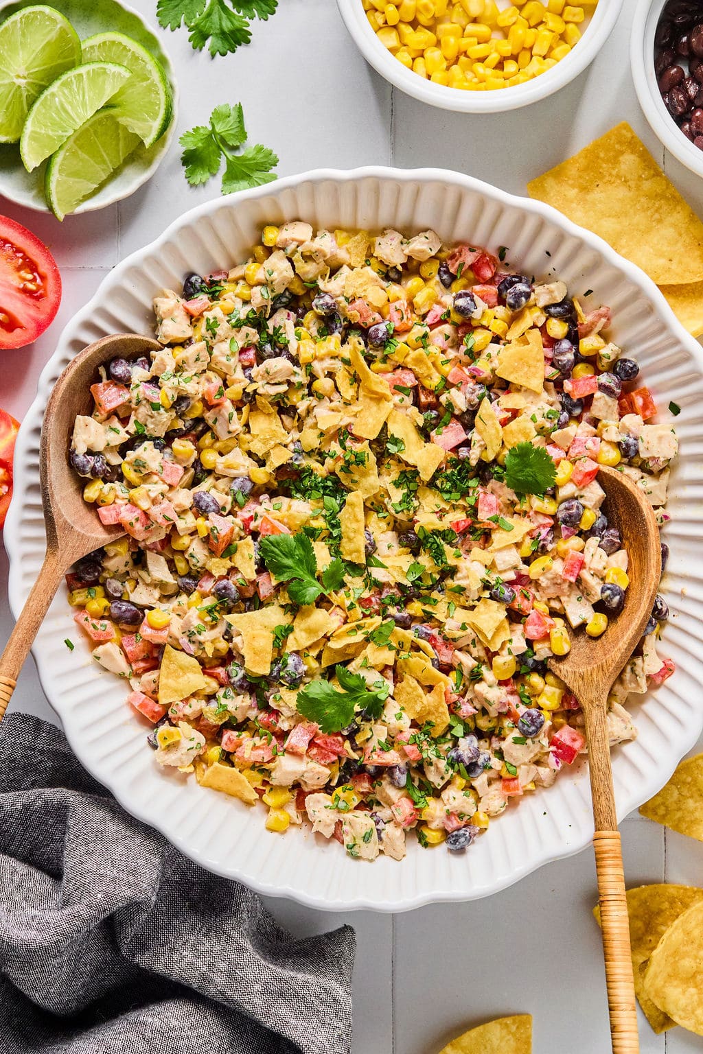 A bowl of southwest-style salad with chopped vegetables, black beans, corn, crushed tortilla chips, and herbs, with two wooden serving spoons.