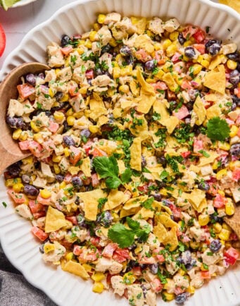 A white dish filled with a mixed salad of corn, black beans, diced vegetables, chicken, topped with crushed tortilla chips and garnished with cilantro.