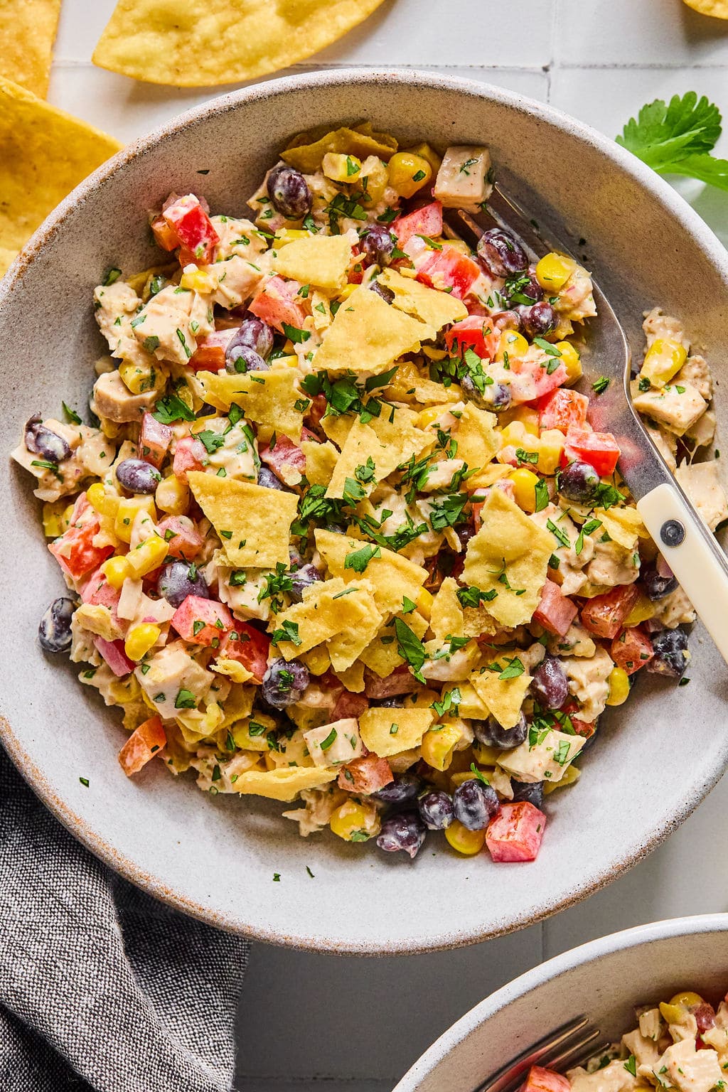 A bowl of creamy salad with black beans, corn, tomatoes, diced chicken, and crushed tortilla chips, garnished with fresh herbs, with a fork resting inside the bowl.