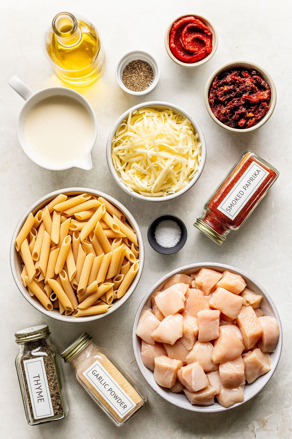 Ingredients for a pasta dish including penne, cubed chicken, shredded cheese, tomato paste, sun-dried tomatoes, oil, cream, and seasonings like smoked paprika, thyme, and garlic powder.