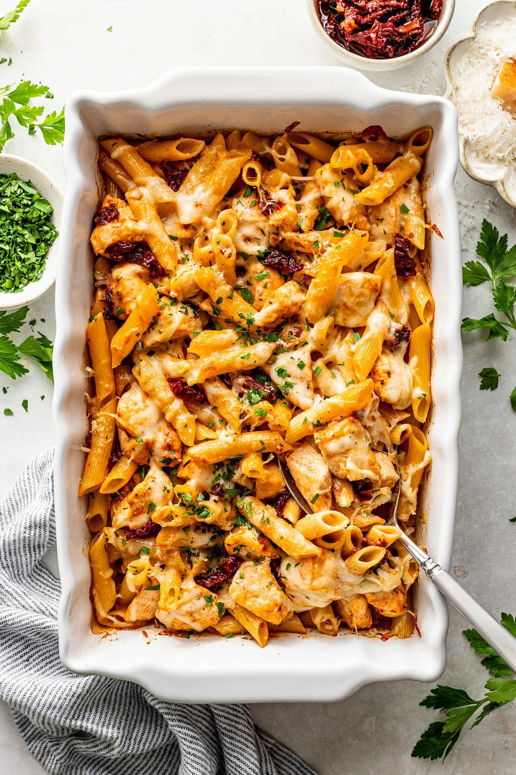 Baked penne pasta with chicken, melted cheese, and sun-dried tomatoes in a white casserole dish with a serving spoon.