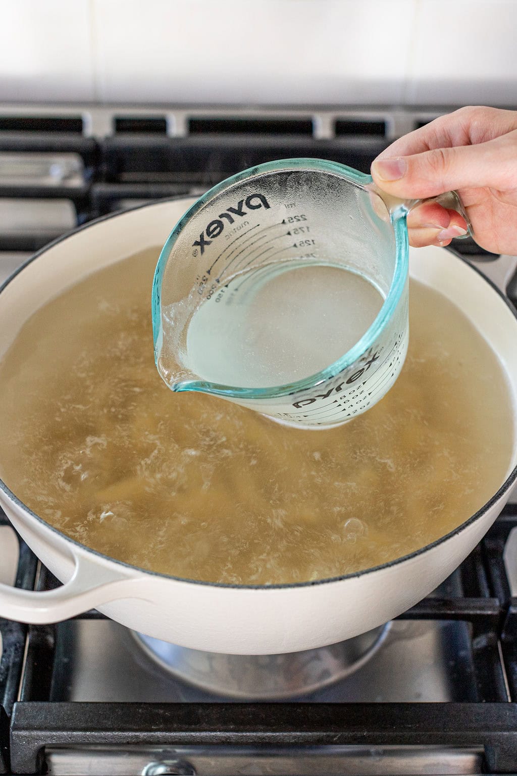 A hand pours liquid from a glass measuring cup into a large pot of boiling water on a stove.