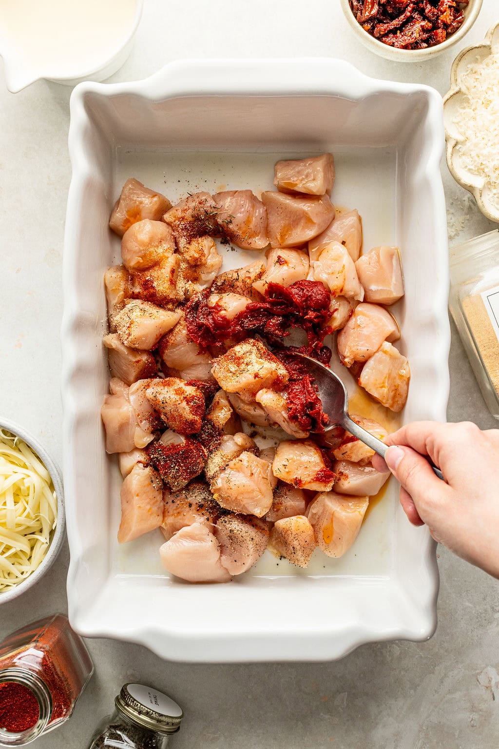 A hand uses a spoon to stir tomato paste and spices into cubed raw chicken in a white baking dish.