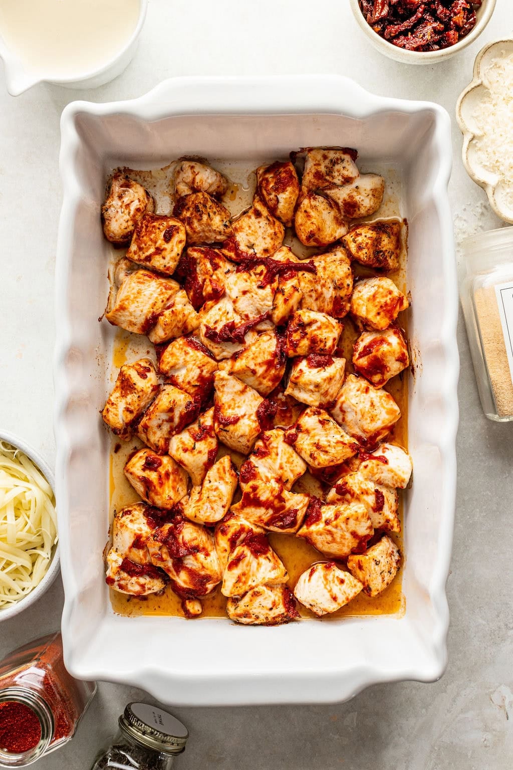 Bite-sized pieces of chicken coated in a red sauce in a white baking dish, surrounded by small bowls of cheese, sun-dried tomatoes, and spices.
