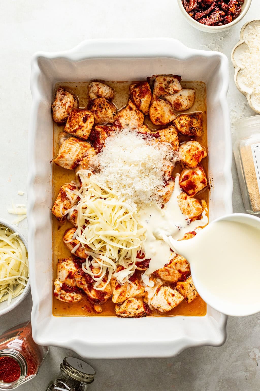 Cream being poured into a baking dish filled with seasoned chicken, shredded cheese, and grated Parmesan.