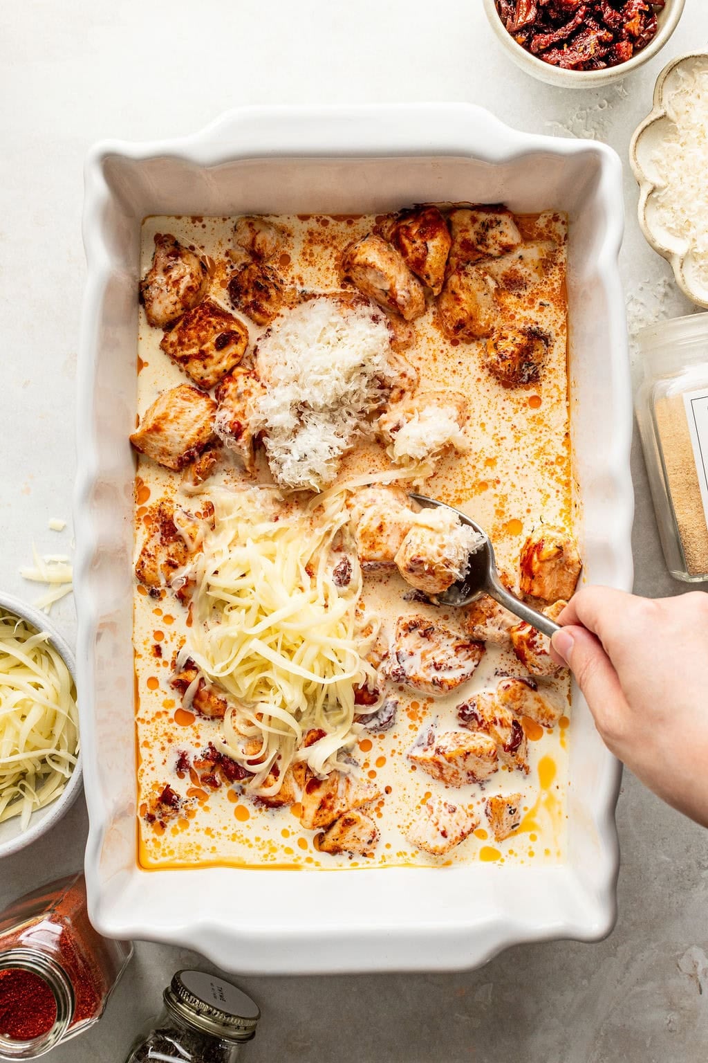 A hand stirs shredded cheese and cream sauce into a baking dish containing cooked chicken pieces and sun-dried tomatoes.