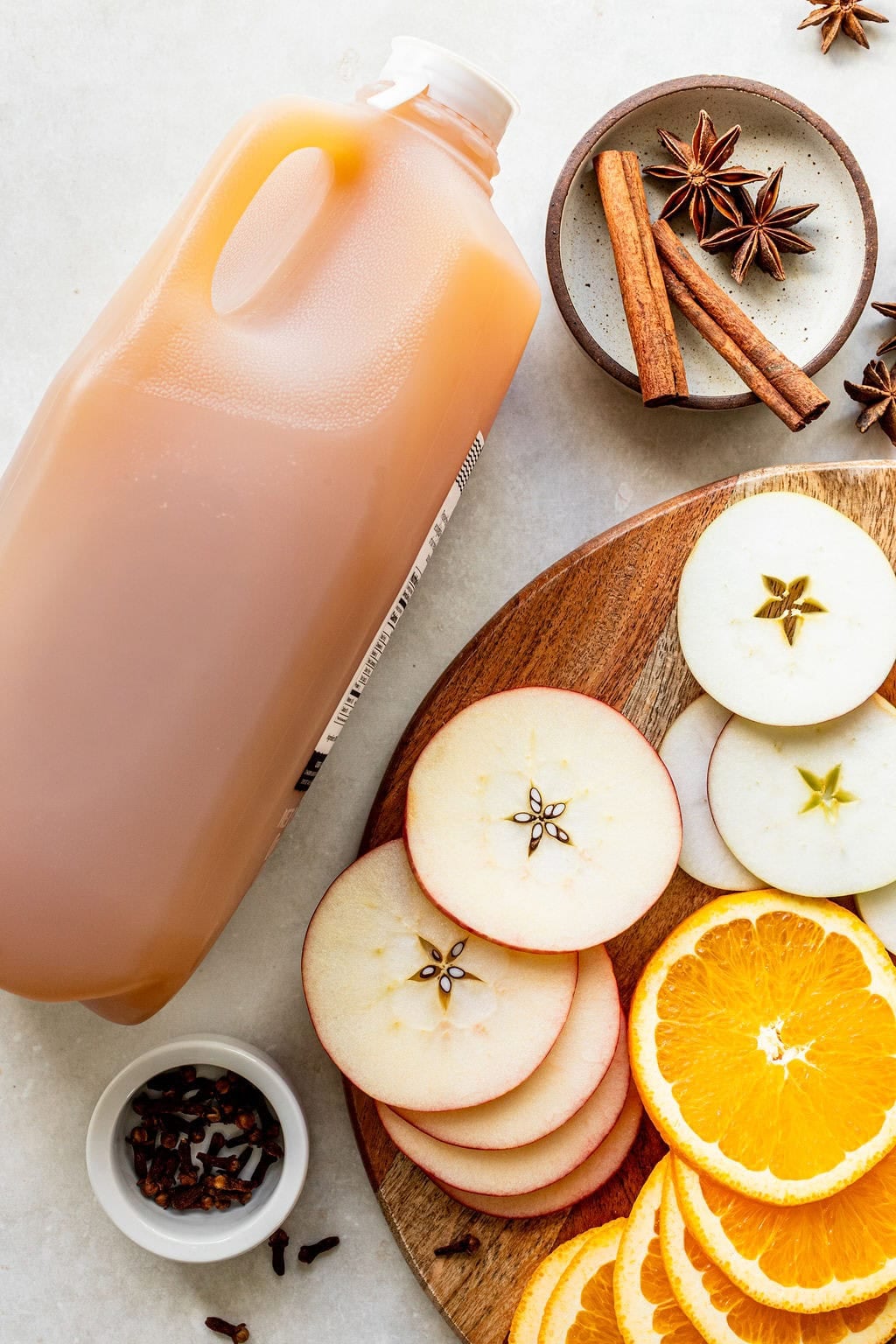 A plastic jug of apple cider next to a wooden board with sliced apples, orange slices, cinnamon sticks, star anise, and whole cloves.