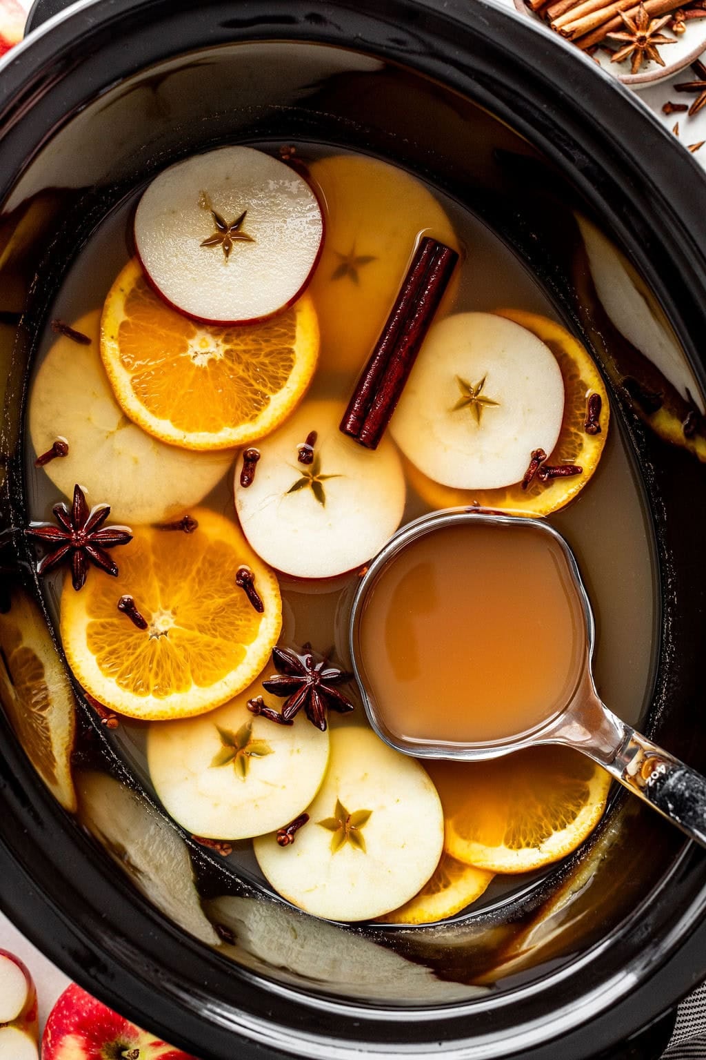 A slow cooker filled with apple cider, garnished with sliced apples, orange wheels, cinnamon sticks, star anise, and cloves, with a metal ladle submerged inside.