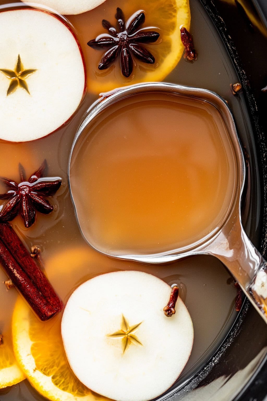 A ladle in a slow cooker filled with mulled cider garnished with apple slices, orange rounds, star anise, and cinnamon sticks.