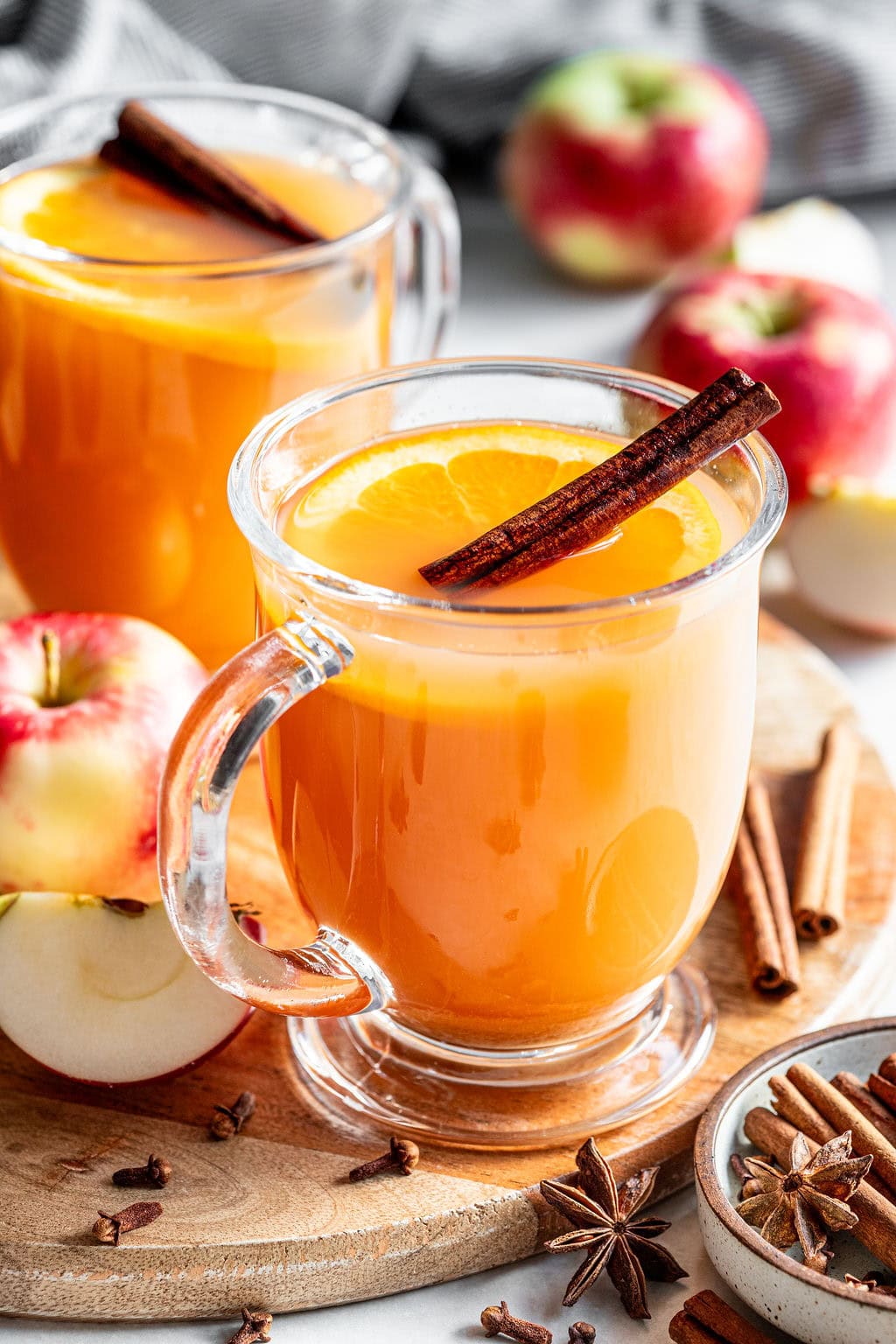 Two glass mugs of apple cider, each garnished with an orange slice and a cinnamon stick, surrounded by fresh apples, star anise, and cloves on a wooden board.