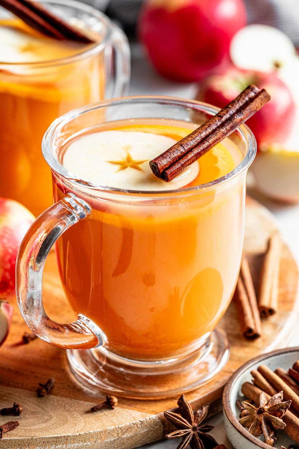 A glass mug of apple cider garnished with an apple slice and a cinnamon stick, with whole apples and spices on a wooden board.