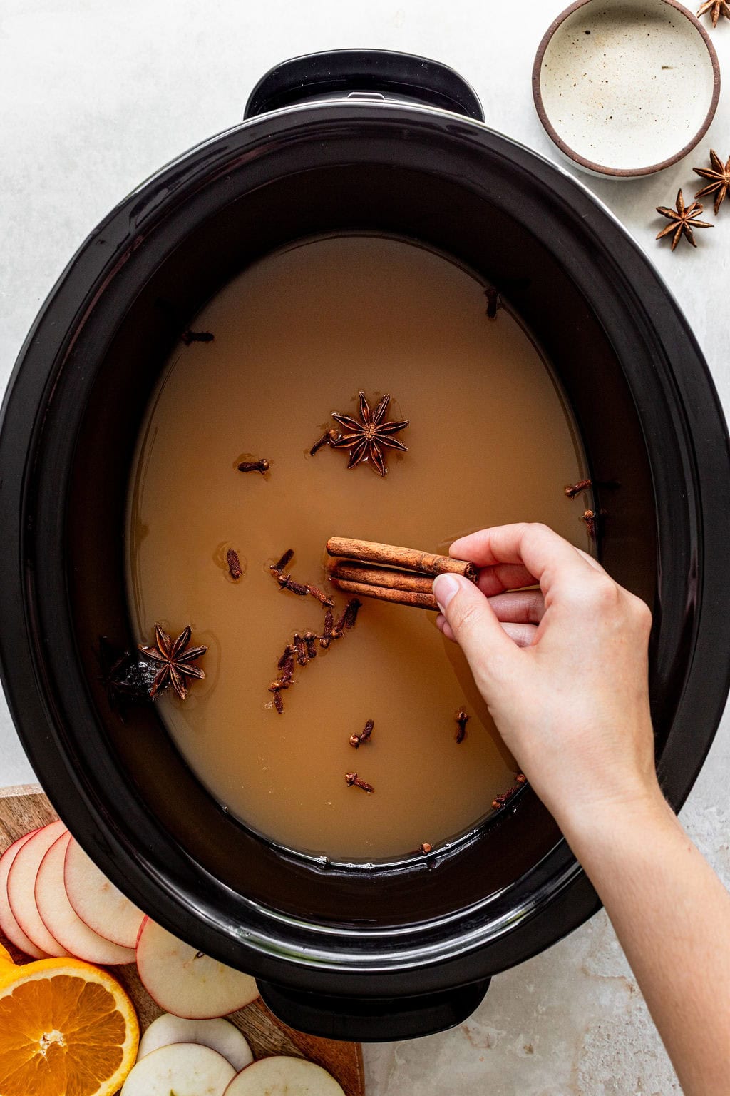 A hand placing cinnamon sticks into a slow cooker filled with liquid, star anise, and cloves.