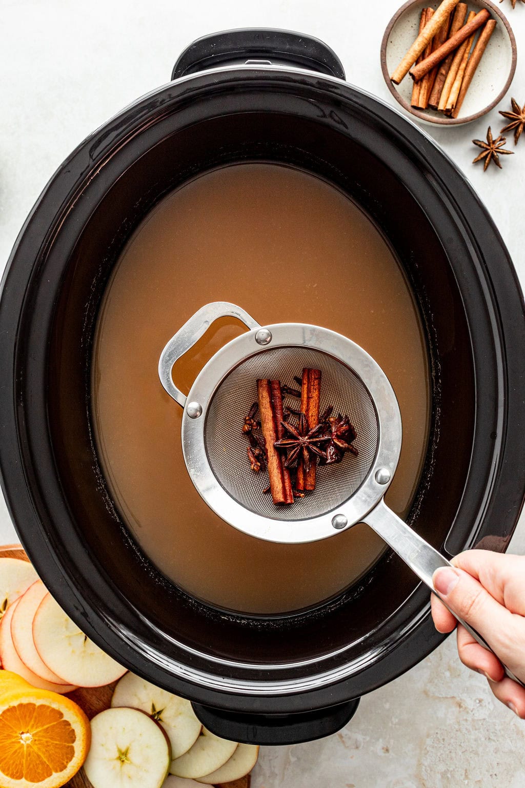 A hand holding a mesh strainer containing cinnamon sticks and star anise over a slow cooker filled with apple cider.
