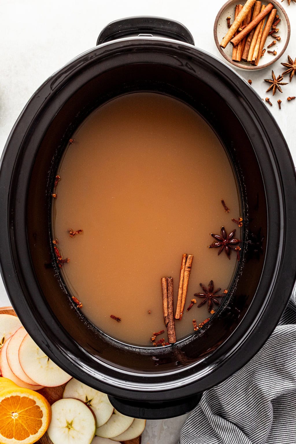 A slow cooker filled with apple cider, spiced with cinnamon sticks, star anise, and cloves, with sliced apples and oranges nearby.
