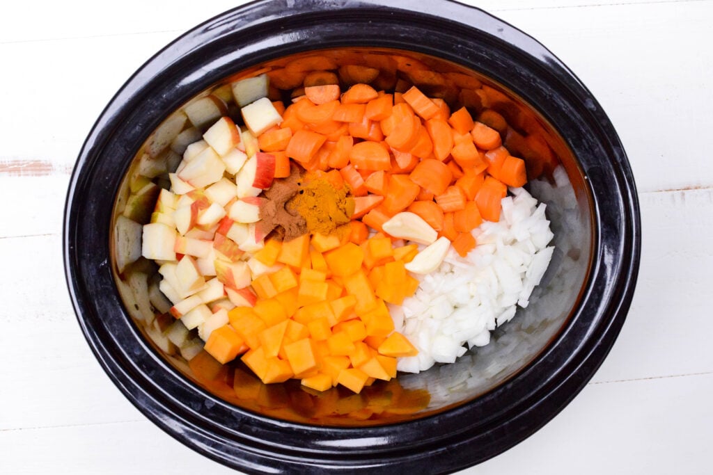 Slow cooker filled with diced apples, butternut squash, onions, garlic cloves, and two mounds of spices.